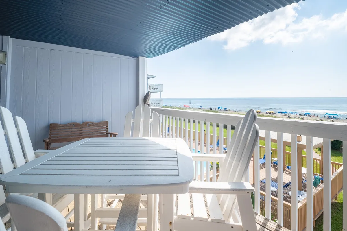 This image showcases a private balcony furnished with white outdoor furniture, offering unobstructed views of the Atlantic Ocean under a bright blue sky. The scene is tranquil, with an inviting table set for dining or relaxing while soaking in the ocean breeze.