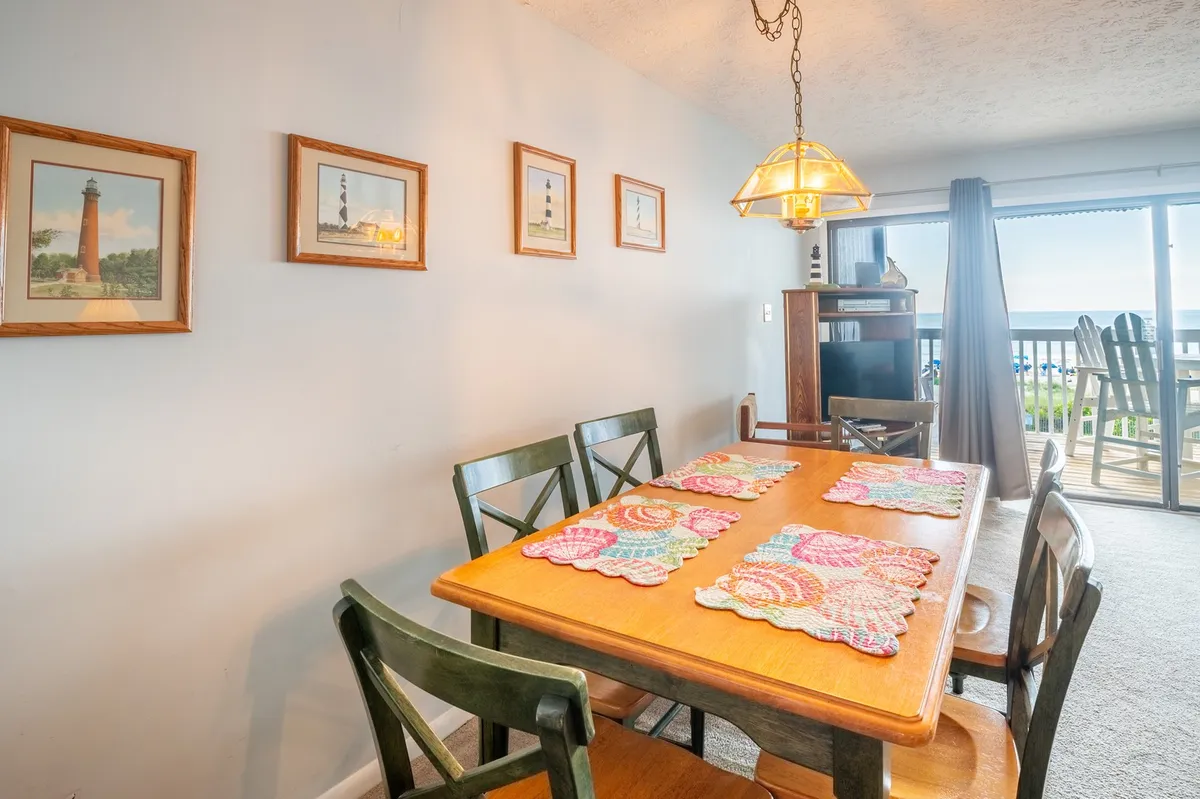 A sunlit dining area featuring a table with colorful placemats, lighthouse pictures adorning the walls, and a window that offers a glimpse outside. The inviting setup suggests a space perfect for casual gatherings and enjoying meals while soaking up the coastal ambiance.