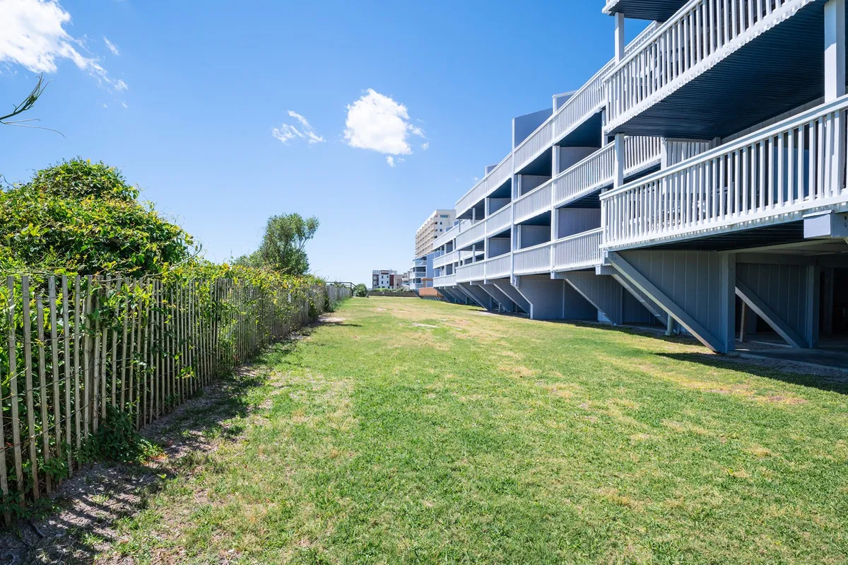A wide, green lawn stretches out in front of the condominium building, surrounded by lush natural greenery. The scene is set for community gatherings, outdoor picnics, or simply enjoying the open space for leisure activities. Balconies overlook the area, tying the outdoor amenities to the residences.