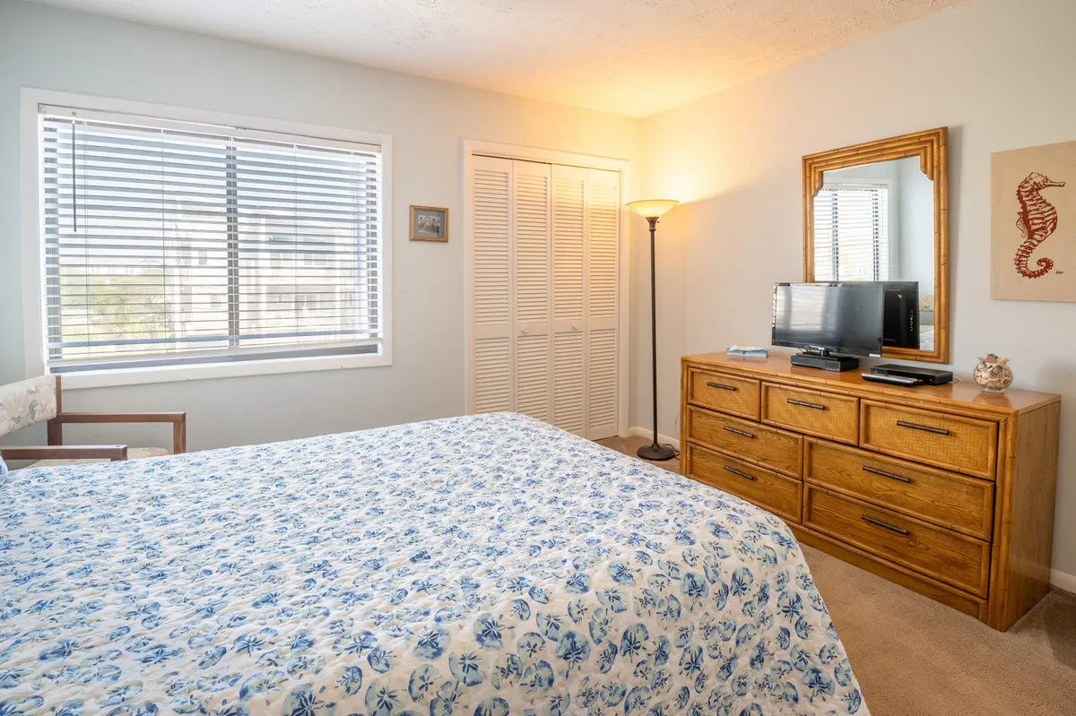 A serene bedroom featuring a queen bed adorned with floral bedding, a wooden dresser, a table lamp, and a window with blinds that lets in soft natural light. There’s also a television placed for easy viewing from the bed, making this room feel cozy yet functional.