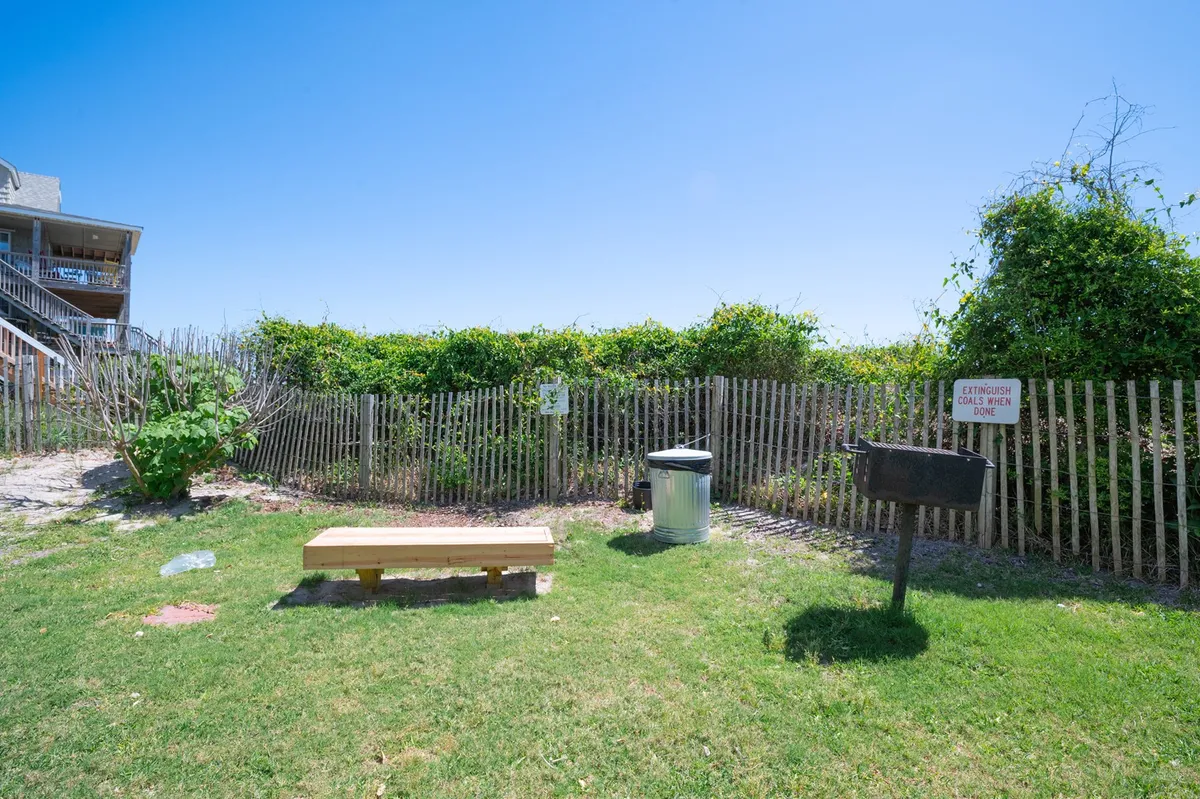 A cozy picnic area featuring a wooden bench, a grill for barbecuing, and a nearby fence. The spot is shaded and inviting, perfect for outdoor meals, casual socializing, or simply relaxing after a day at the beach.