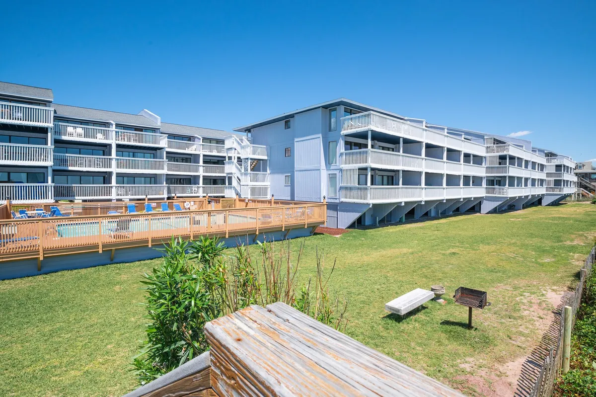 This exterior image captures the oceanfront pool surrounded by balconies, green lawns, and a barbecue area. The pool is the focal point, with families and friends enjoying swimming and sunbathing. The green space and barbecue facilities convey an inviting atmosphere for outdoor gatherings.