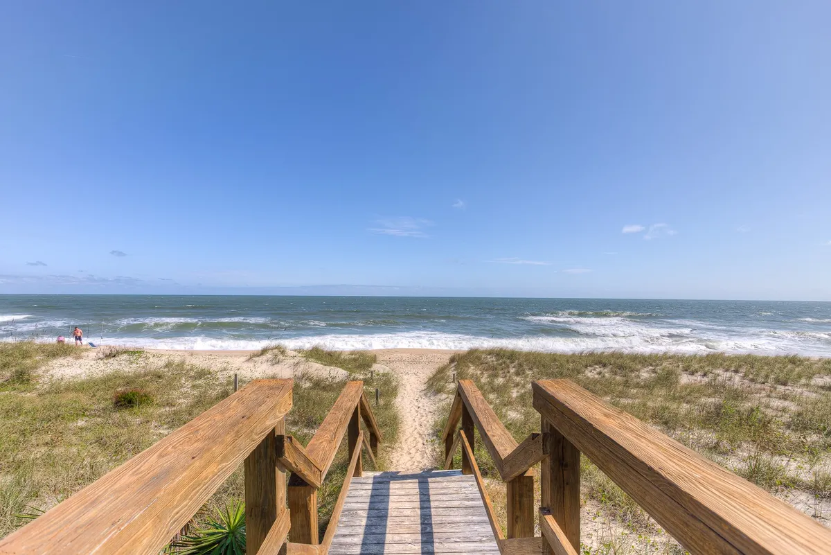 A scenic shot of a wooden walkway leading directly to the sandy beach, with ocean waves and dunes in view under a serene sky. This image evokes the anticipation of a day by the sea, as seen from the vantage point of a beachfront property.