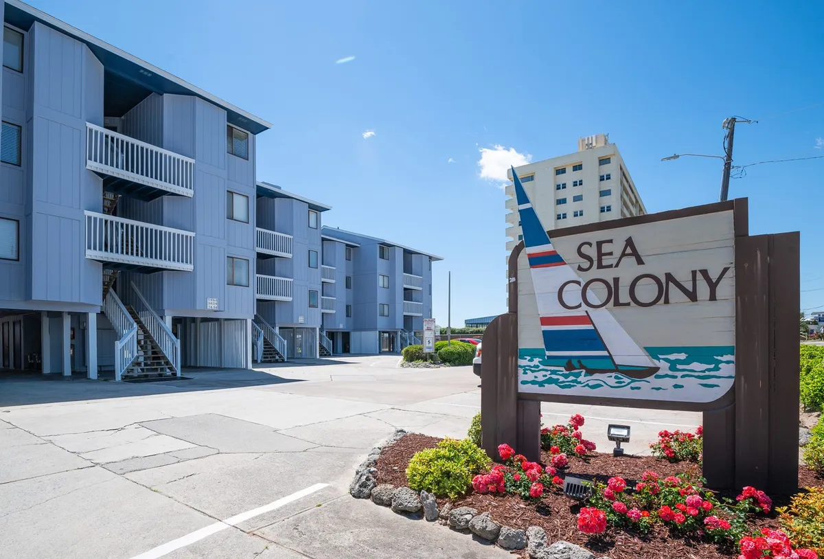 The exterior of Sea Colony is highlighted, featuring a welcoming blue and gray facade, a vibrant flower bed with red blooms, and the prominent Sea Colony sign. The image hints at its proximity to the beach and the relaxed, inviting ambiance of the community.