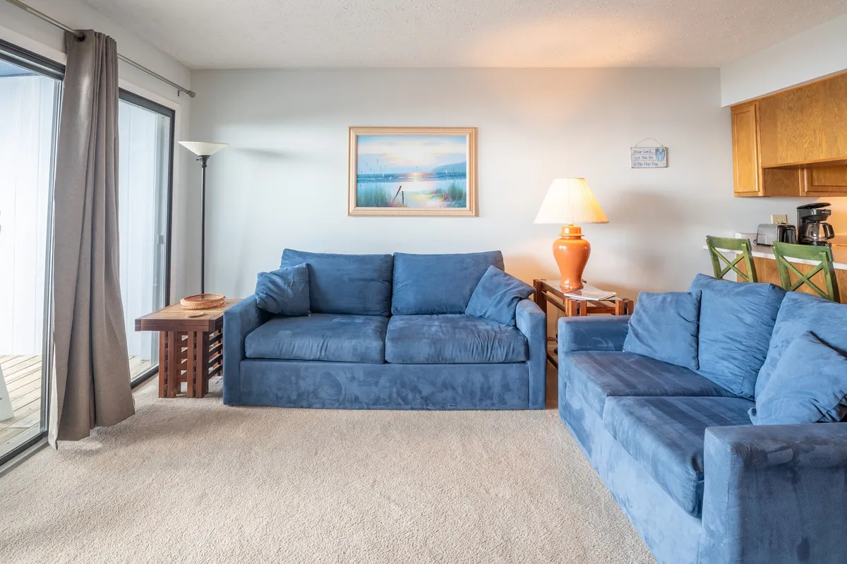 A welcoming interior scene with blue sofas, a lamp, wall art, and a visible kitchen area in the background. The layout supports socializing and family activities, capturing both relaxation and meal prep zones.