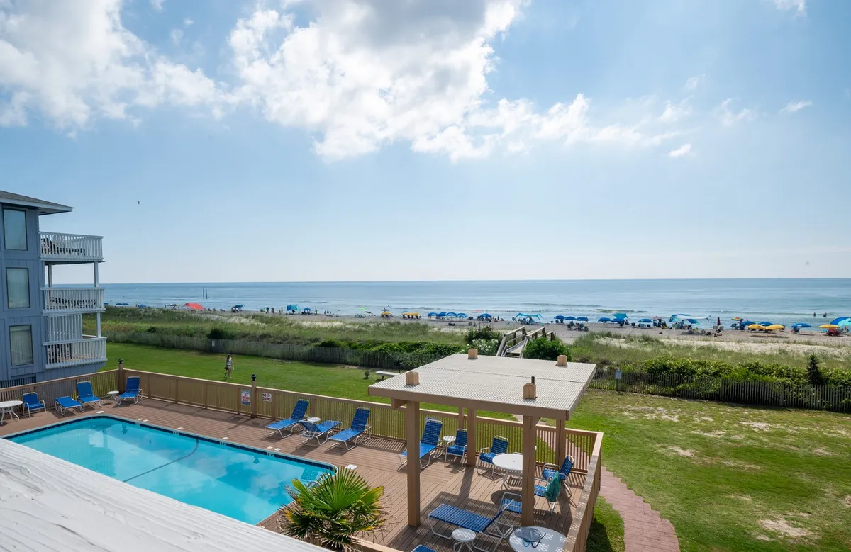 An exterior view from the deck area, featuring the ocean in the background, a swimming pool, deck chairs, and beach umbrellas. The scene captures the transition from the building to the balcony and out to the ocean beyond, with a sense of leisure and coastal atmosphere.