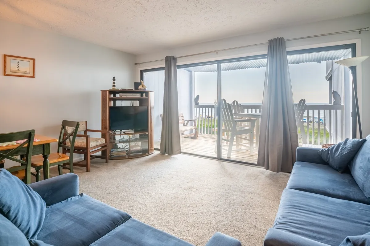 A cozy open-plan living and dining area featuring a comfortable sofa, a dining table with chairs, a television, and a wall of sliding glass doors that open to ocean views. The space is filled with natural light and has direct access to a balcony.