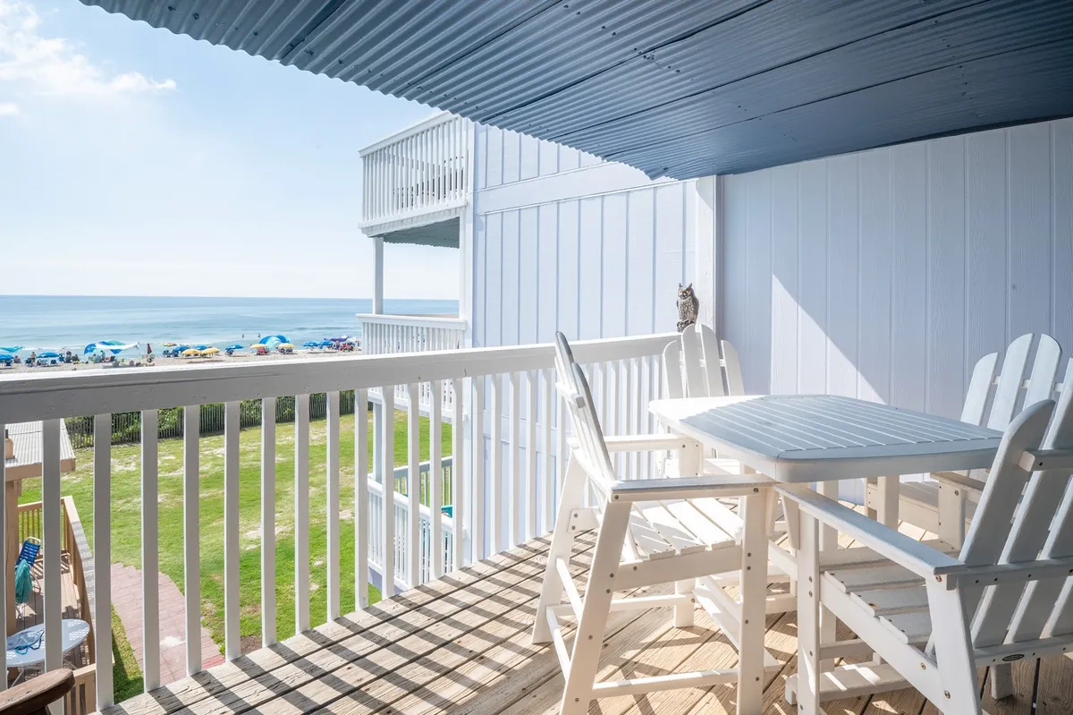 The image showcases a tranquil oceanfront balcony with inviting outdoor furniture and bright beach umbrellas, all overlooking the sparkling Atlantic. The setting is perfect for relaxing mornings watching the sunrise or gathering in the evening for breezy, starlit conversations, with the sand and surf just steps away.