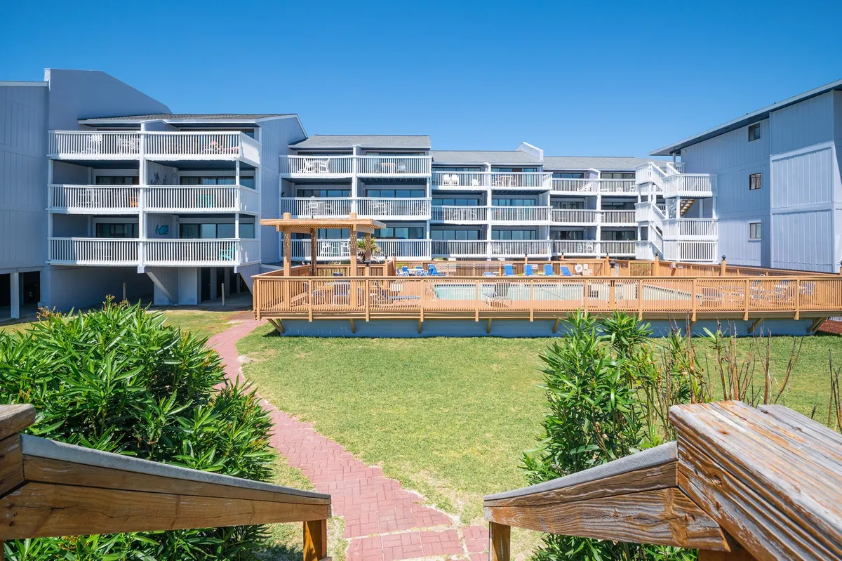 A walkway leads past a swimming pool and lush landscaping towards balconies that offer elevated views. The image suggests easy movement between the building amenities and outdoor spaces, hinting at convenient access to both the pool and the beach.