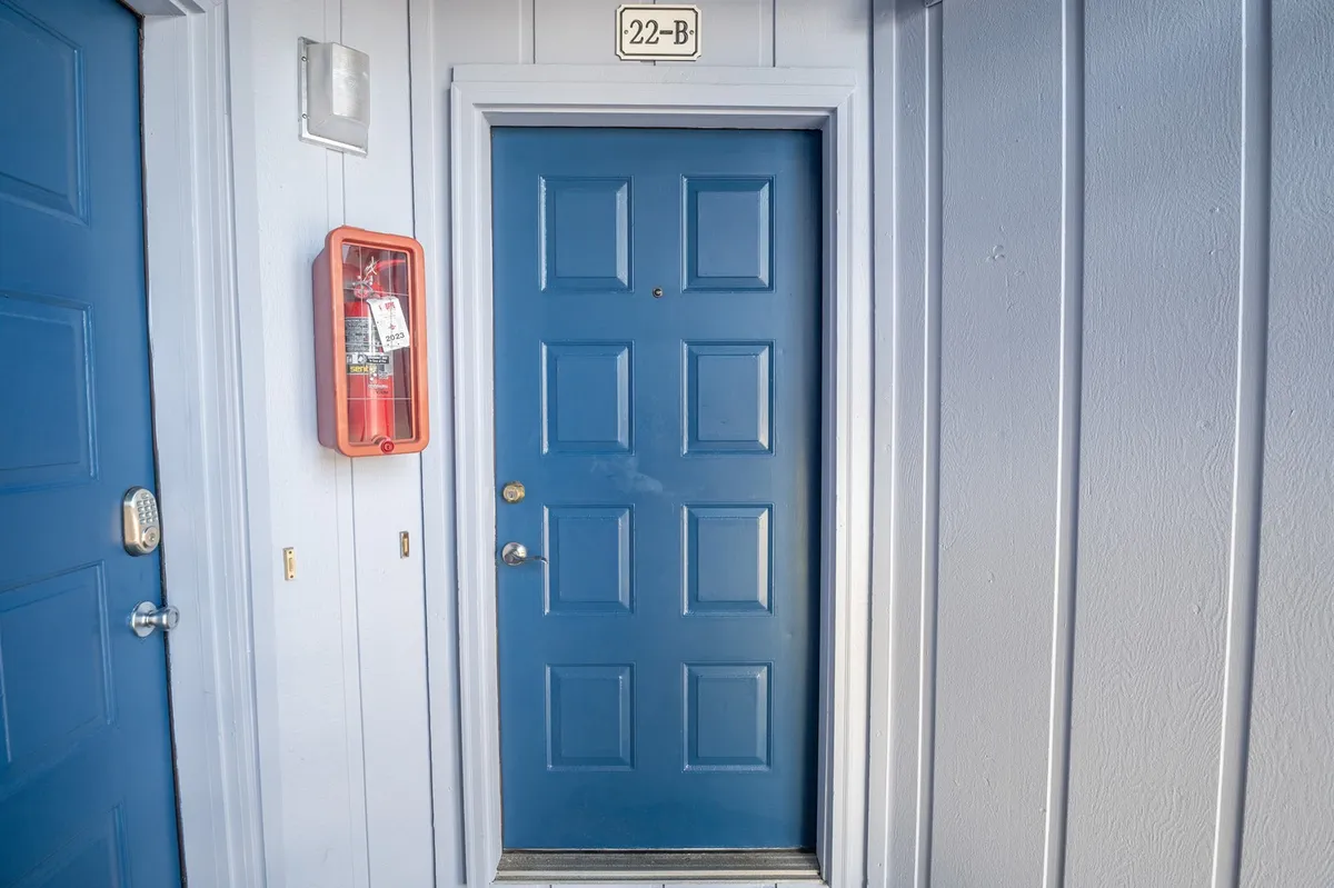 This image features the blue front door of a unit, clearly labeled with the door number 22-B and a fire alarm box nearby. The scene suggests an inviting entrance, with the door standing as a threshold between the private accommodations and the surrounding community.