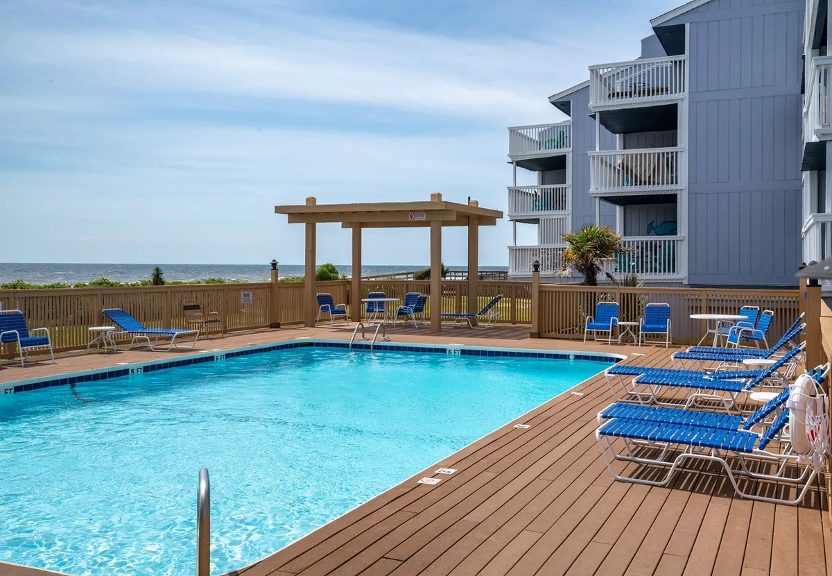 A sparkling swimming pool with blue lounge chairs and a canopy structure overlooks the ocean in the background. The setup invites relaxation and socializing, while the ocean view beyond the pool connects directly to the beachside experience.